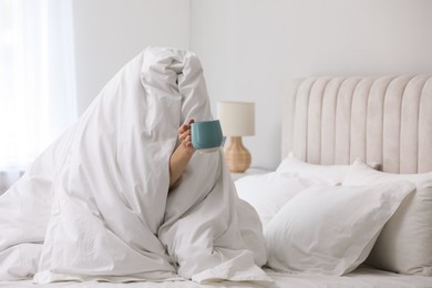 Photo of Woman wrapping in blanket sitting with cup of coffee on bed. Good morning