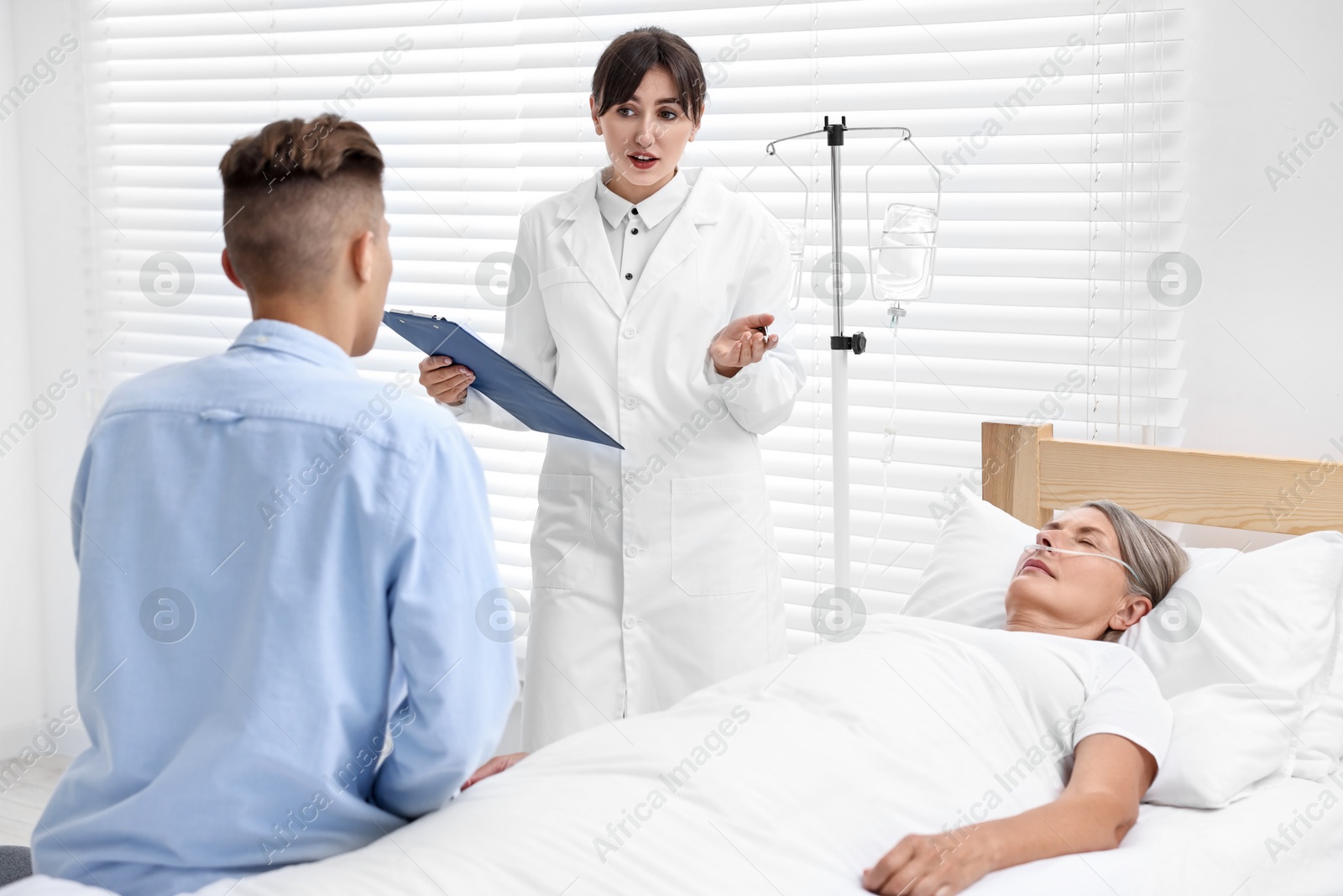 Photo of Coma patient. Sad young man near his unconscious mother talking with doctor in hospital