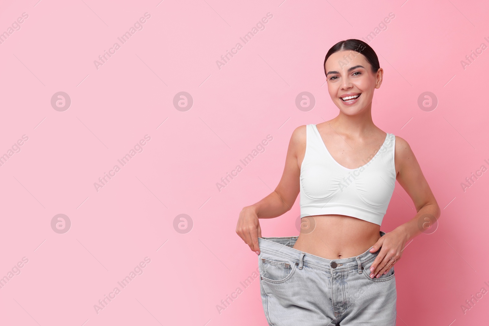 Photo of Diet and weight loss concept. Happy young woman in big jeans showing her slim body on pink background. Space for text