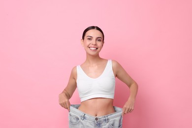 Photo of Diet and weight loss concept. Happy young woman in big jeans showing her slim body on pink background