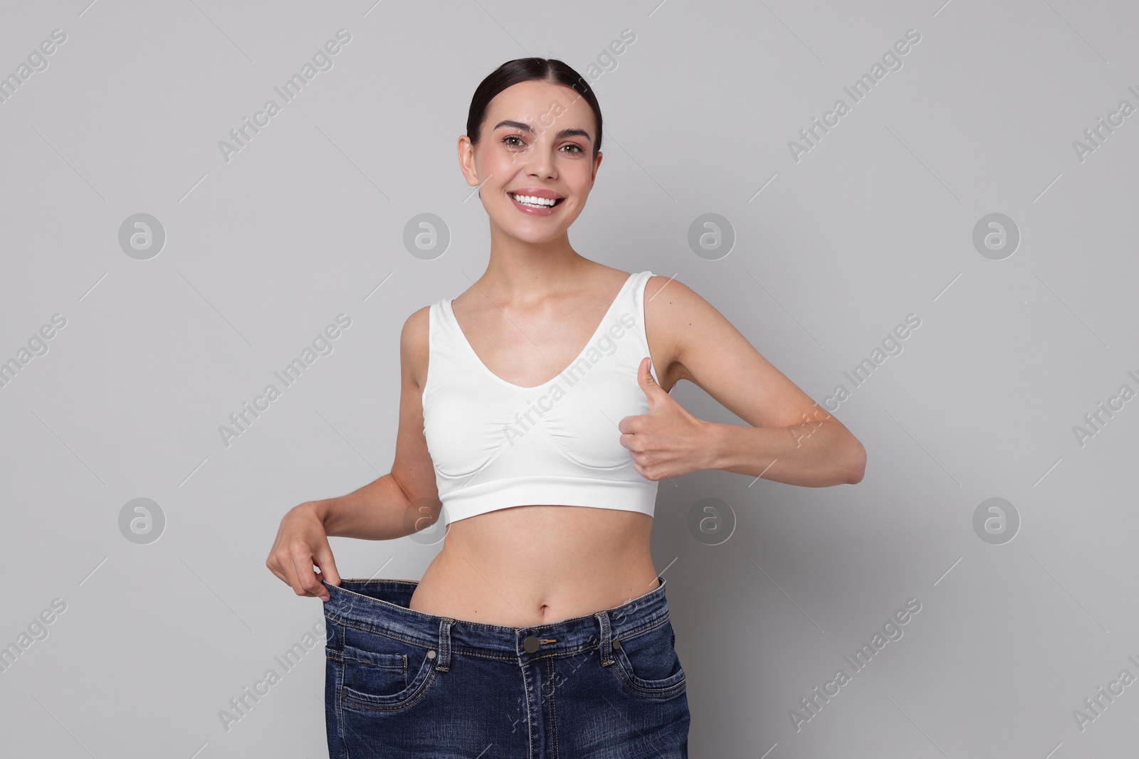 Photo of Diet and weight loss concept. Happy young woman in big jeans showing her slim body on light grey background