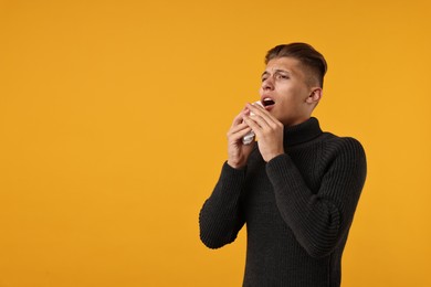 Photo of Cold symptoms. Sick man with tissue sneezing on orange background, space for text