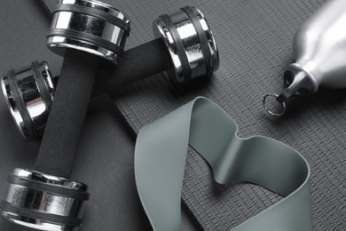 Dumbbells, yoga mat, thermo bottle and fitness elastic band on black background, above view