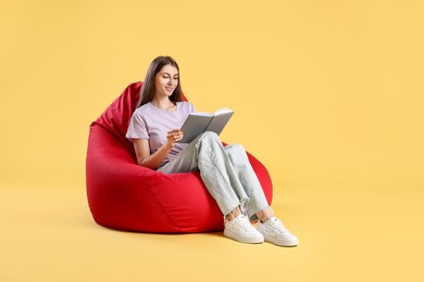 Photo of Smiling woman with book sitting on red bean bag chair against yellow background, space for text