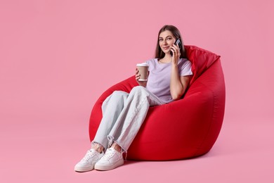 Photo of Beautiful young woman with paper cup of drink talking on smartphone while sitting red bean bag chair against pink background