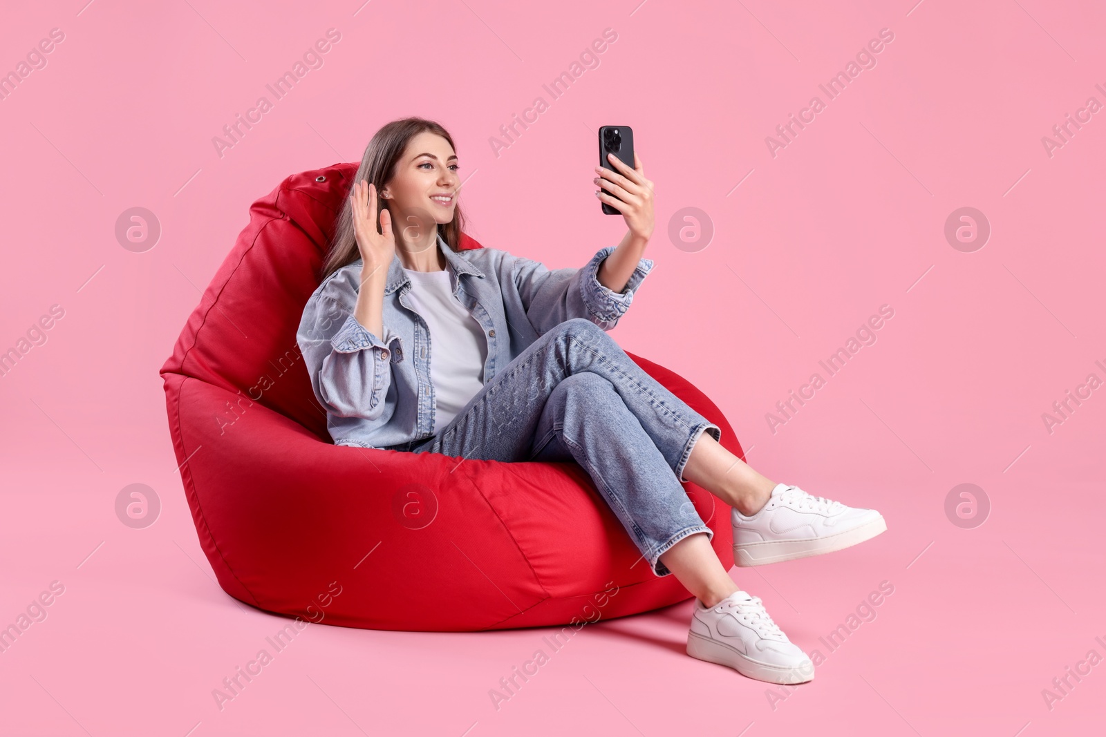 Photo of Smiling woman with smartphone having online meeting on red bean bag chair against pink background