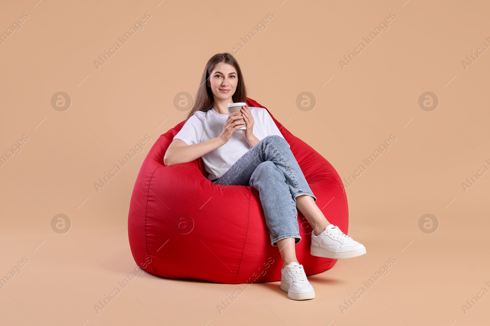 Photo of Beautiful young woman with paper cup of drink on red bean bag chair against beige background