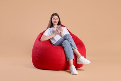 Photo of Beautiful young woman with paper cup of drink on red bean bag chair against beige background