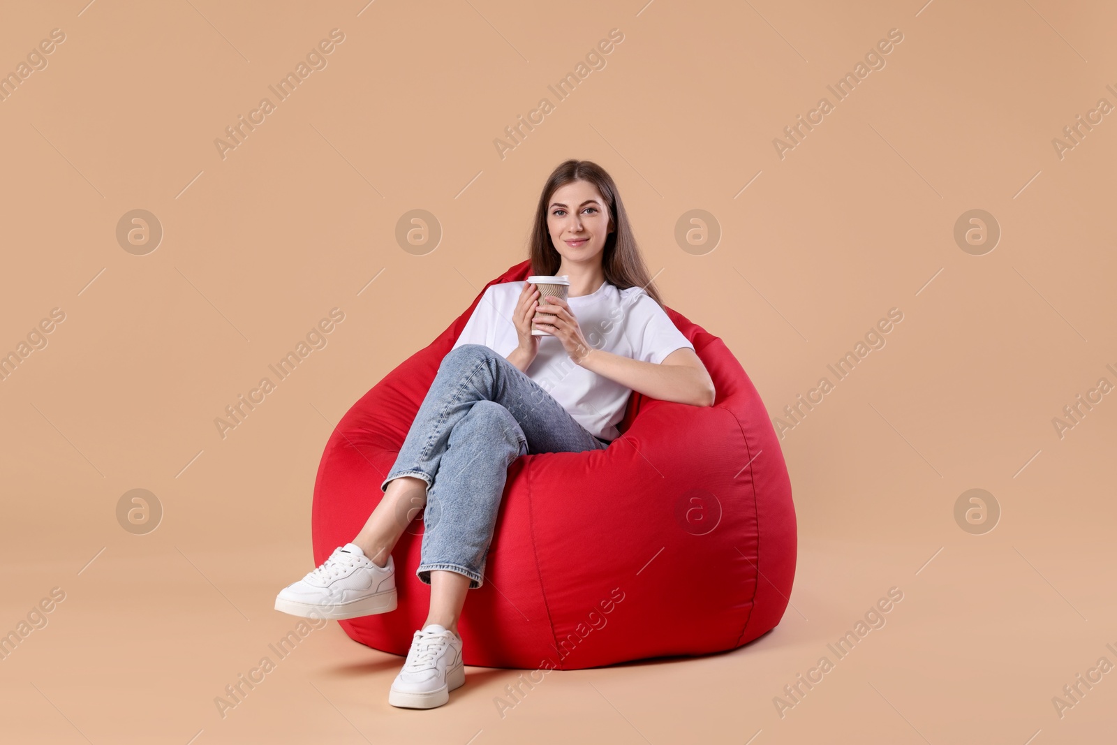 Photo of Beautiful young woman with paper cup of drink on red bean bag chair against beige background