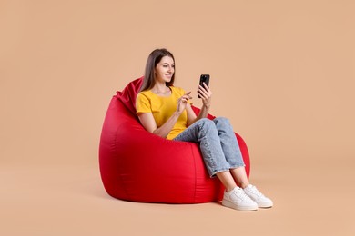 Photo of Smiling woman using smartphone while sitting on red bean bag chair against beige background