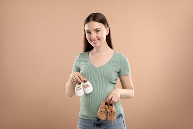 Photo of Expecting twins. Pregnant woman holding two pairs of shoes on light brown background