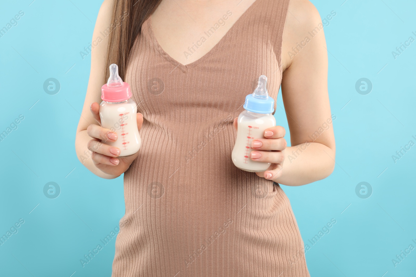 Photo of Expecting twins. Pregnant woman holding two bottles with milk on light blue background, closeup