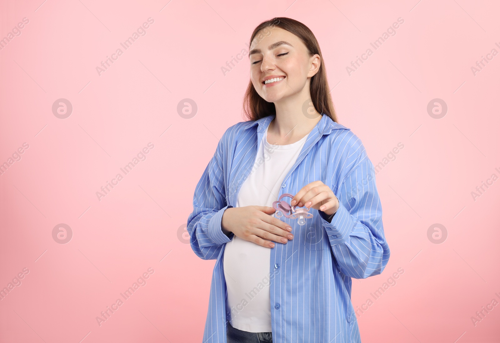 Photo of Expecting twins. Pregnant woman holding two pacifiers on pink background, space for text