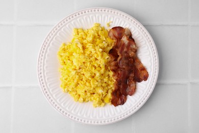 Delicious scrambled eggs with bacon on plate on white tiled table, top view