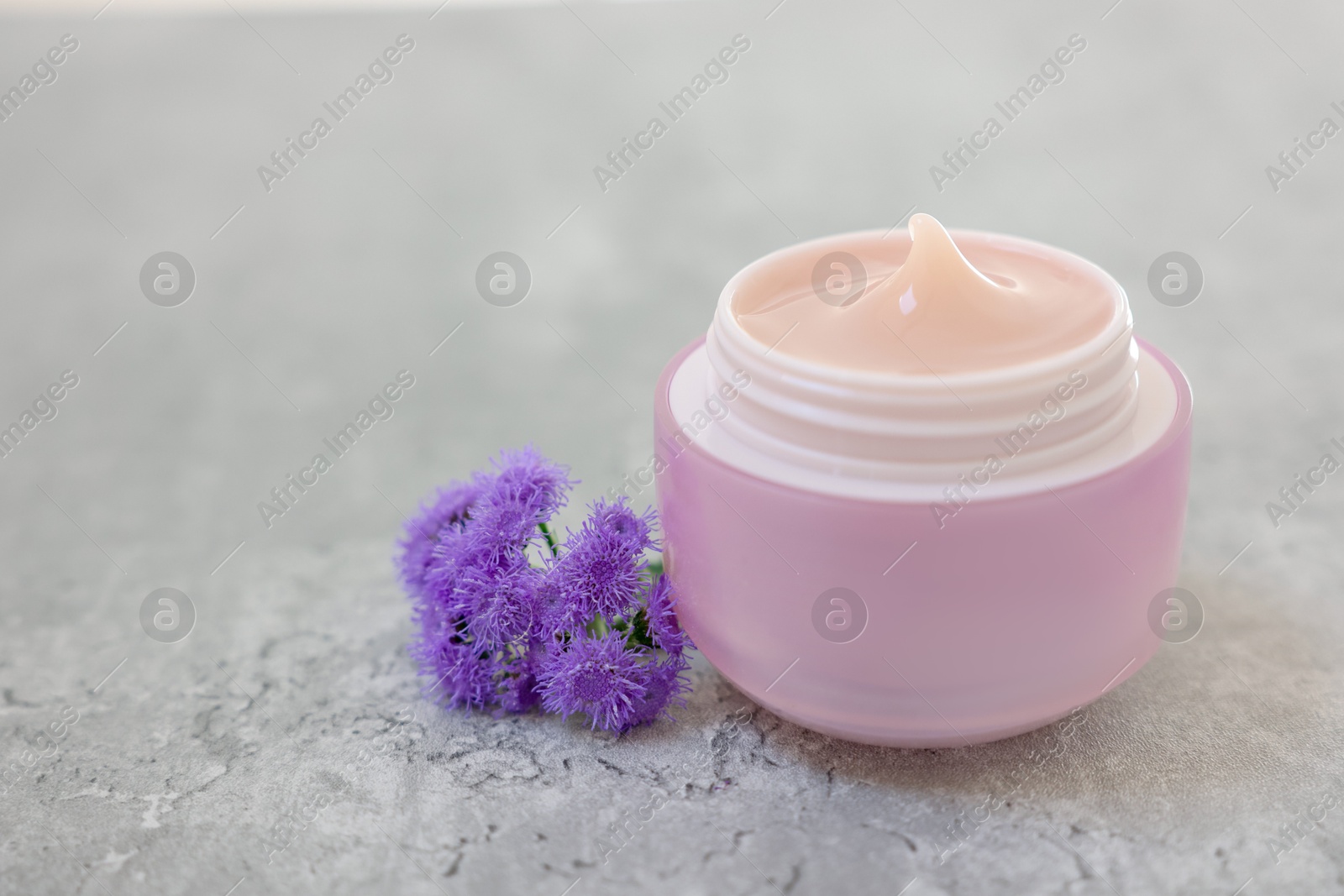 Photo of Jar of cream and ageratum flowers on gray table, closeup. Space for text