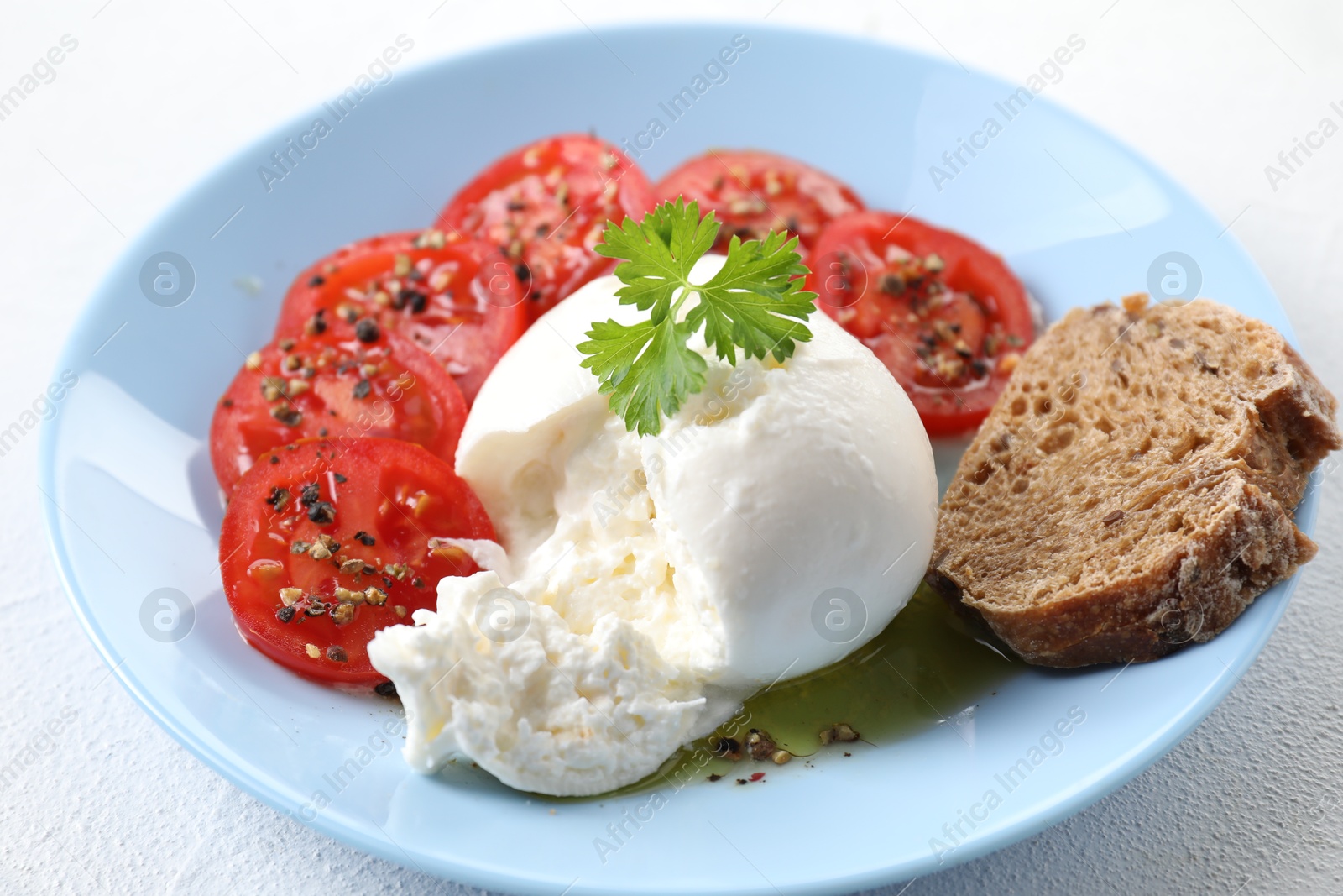 Photo of Delicious burrata cheese, tomatoes, parsley and bread on white table, closeup