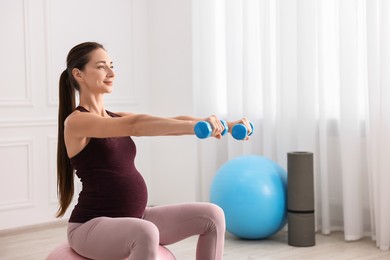 Beautiful pregnant woman with dumbbells doing exercises on fitball at home