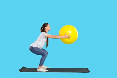 Beautiful pregnant woman doing exercises with fitball on mat against light blue background