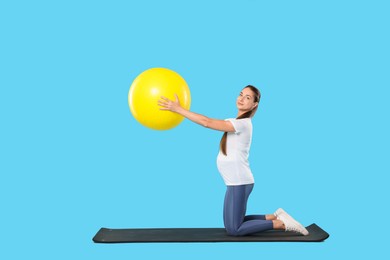 Beautiful pregnant woman doing exercises with fitball on mat against light blue background