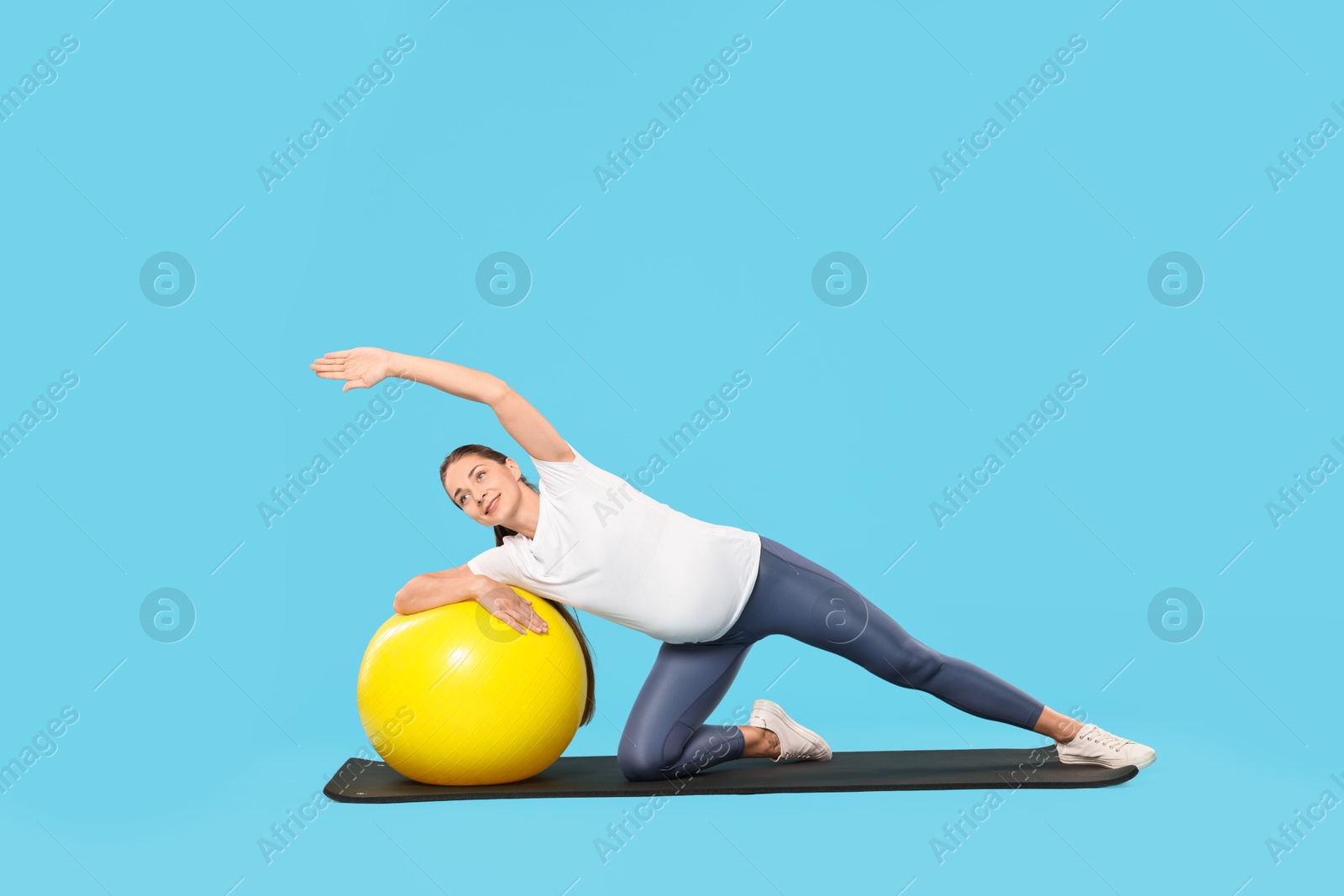 Photo of Beautiful pregnant woman doing exercises with fitball on mat against light blue background