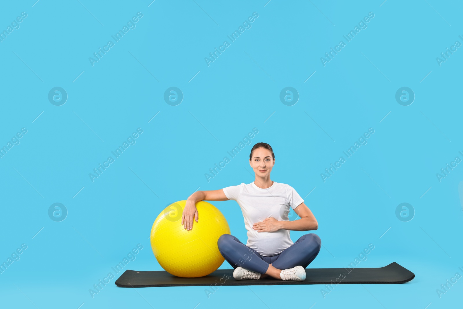 Photo of Beautiful pregnant woman with fitball on exercise mat against light blue background