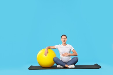 Beautiful pregnant woman with fitball on exercise mat against light blue background