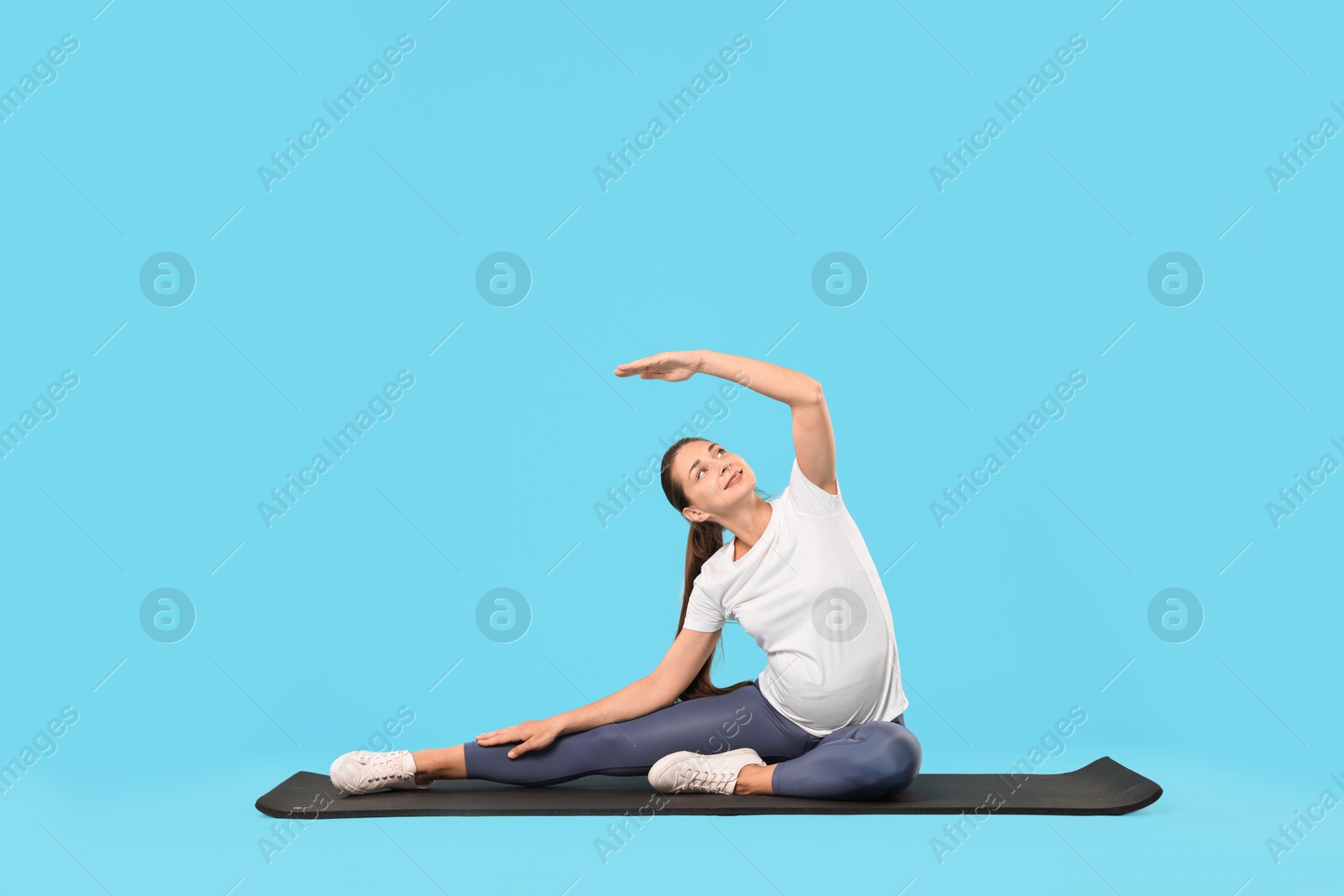 Photo of Beautiful pregnant woman doing stretching exercises on mat against light blue background