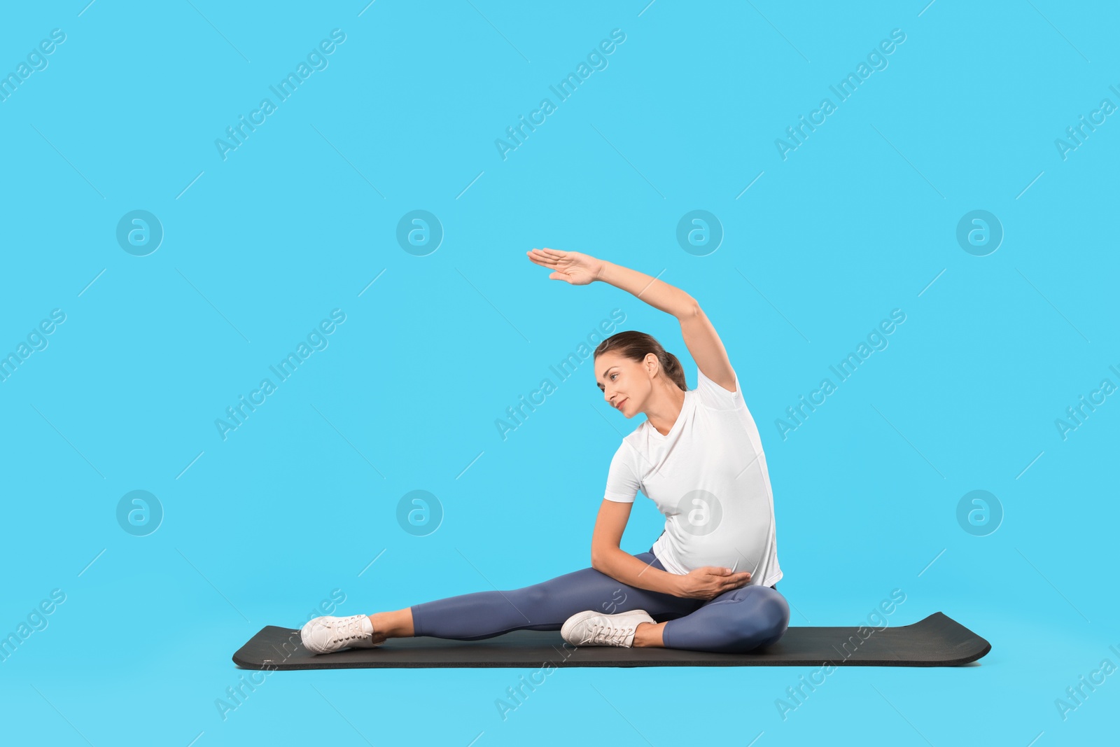 Photo of Beautiful pregnant woman doing stretching exercises on mat against light blue background