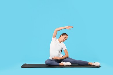 Beautiful pregnant woman doing stretching exercises on mat against light blue background