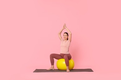 Beautiful pregnant woman doing exercises on fitball against pink background