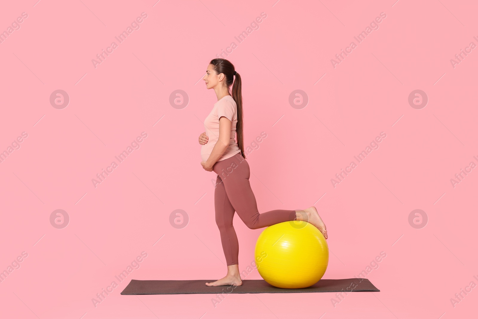 Photo of Beautiful pregnant woman with fitball doing exercises on mat against pink background