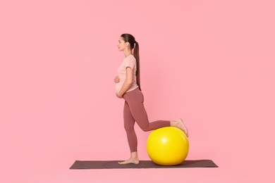 Beautiful pregnant woman with fitball doing exercises on mat against pink background