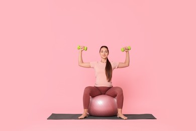 Beautiful pregnant woman with dumbbells doing exercises on fitball against pink background