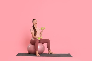 Photo of Beautiful pregnant woman with dumbbells doing exercises on fitball against pink background, space for text