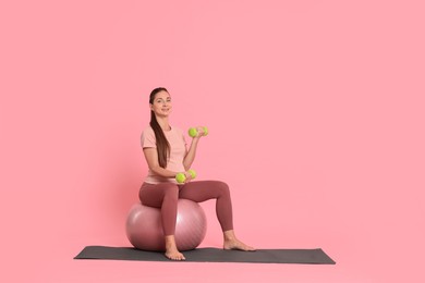 Beautiful pregnant woman with dumbbells doing exercises on fitball against pink background, space for text