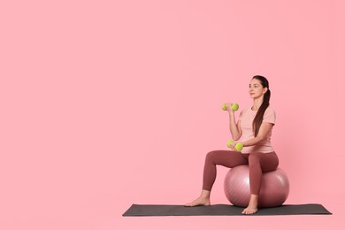 Beautiful pregnant woman with dumbbells doing exercises on fitball against pink background, space for text