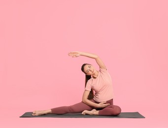 Beautiful pregnant woman doing stretching exercises on mat against pink background