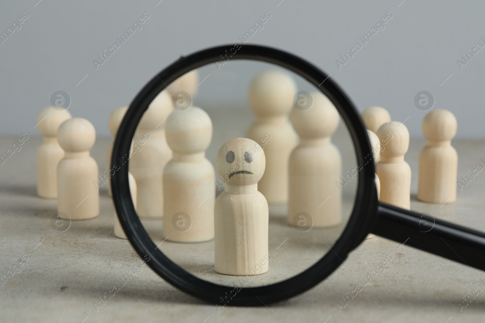 Photo of Magnifying glass showing wooden figure with sad face on grey table, closeup