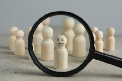 Magnifying glass showing wooden figure with sad face on grey table, closeup