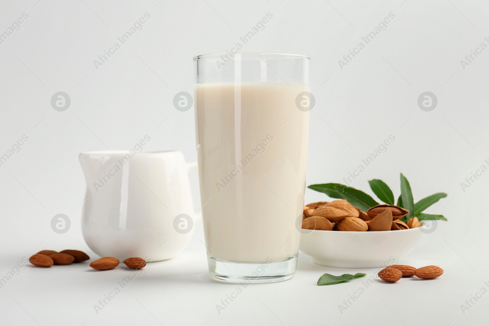 Photo of Fresh almond milk in glass, nuts, green leaves and pitcher on white background