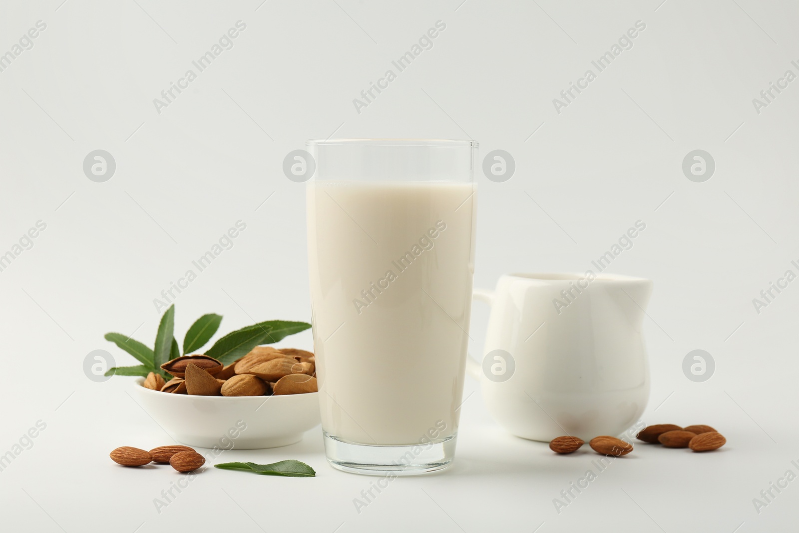 Photo of Fresh almond milk in glass, nuts, green leaves and pitcher on white background