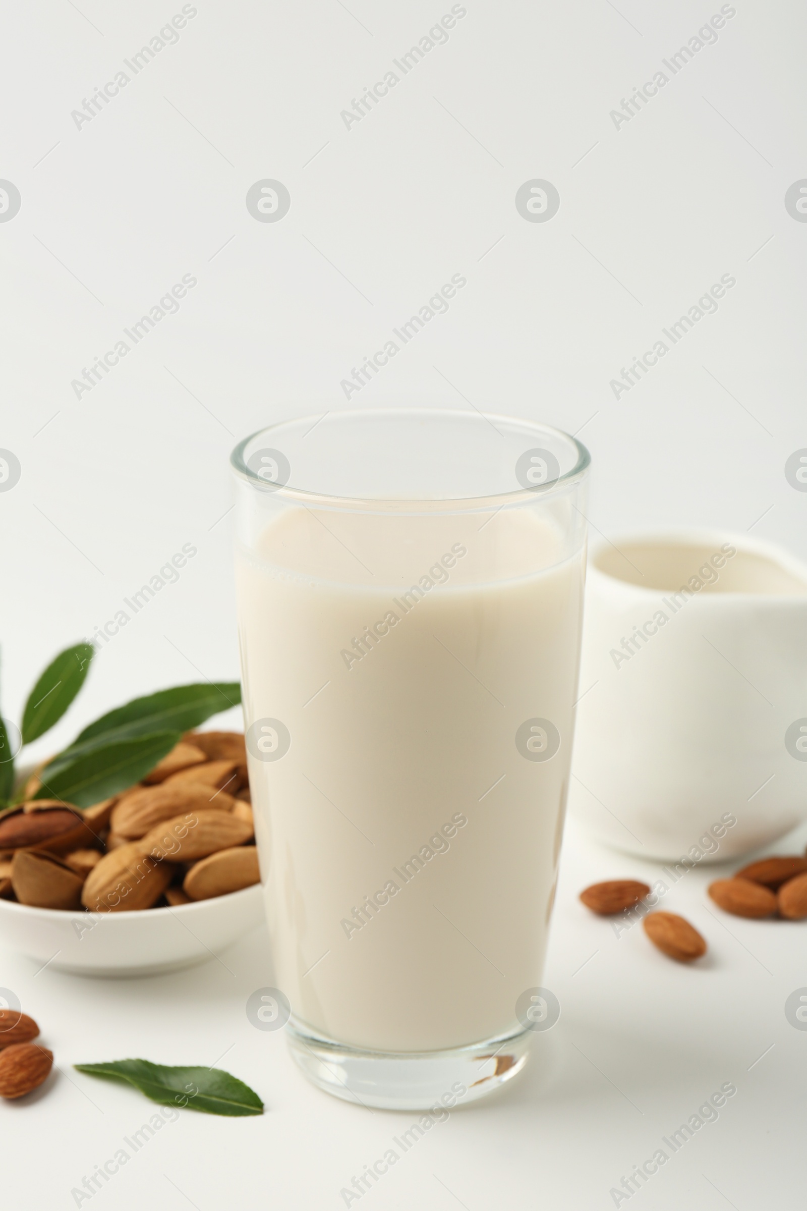 Photo of Fresh almond milk in glass, nuts, green leaves and pitcher on white background