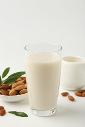 Fresh almond milk in glass, nuts, green leaves and pitcher on white background