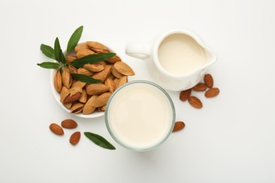 Photo of Fresh almond milk in glass, nuts, green leaves and pitcher on white background, flat lay