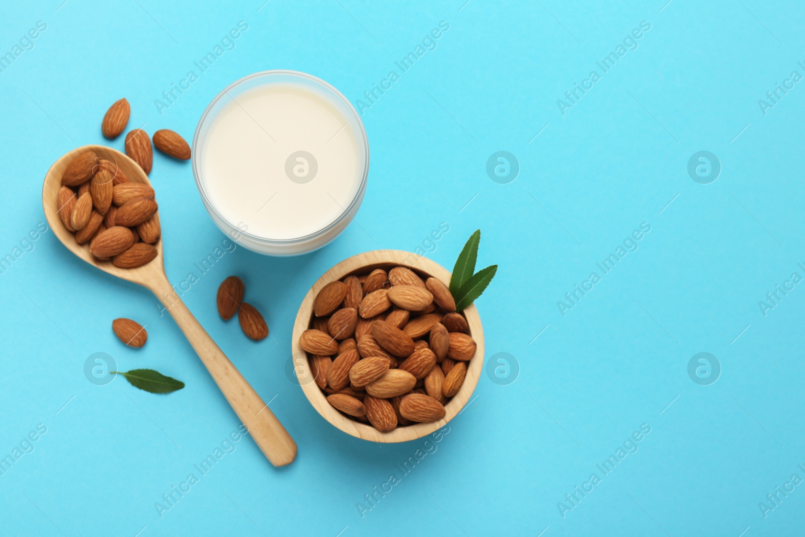 Photo of Fresh almond milk in glass, nuts, spoon and green leaves on light blue background, flat lay. Space for text