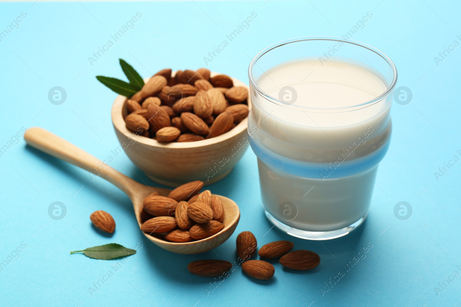 Photo of Fresh almond milk in glass, nuts, spoon and green leaves on light blue background