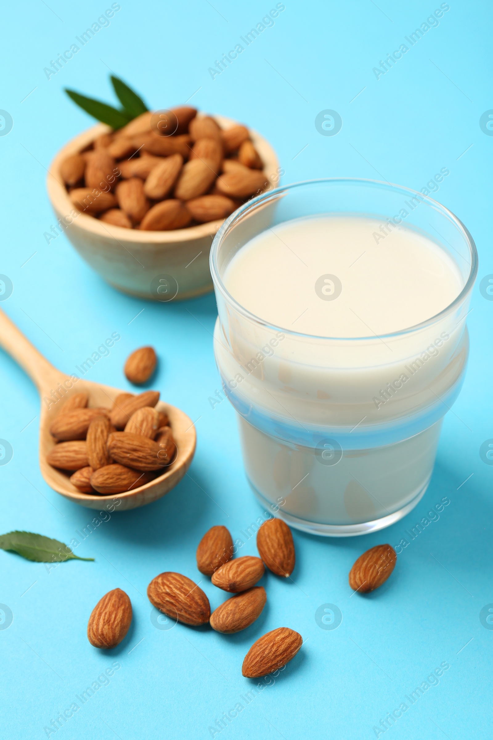 Photo of Fresh almond milk in glass, nuts, spoon and green leaves on light blue background