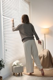 Young woman near window blinds at home, back view