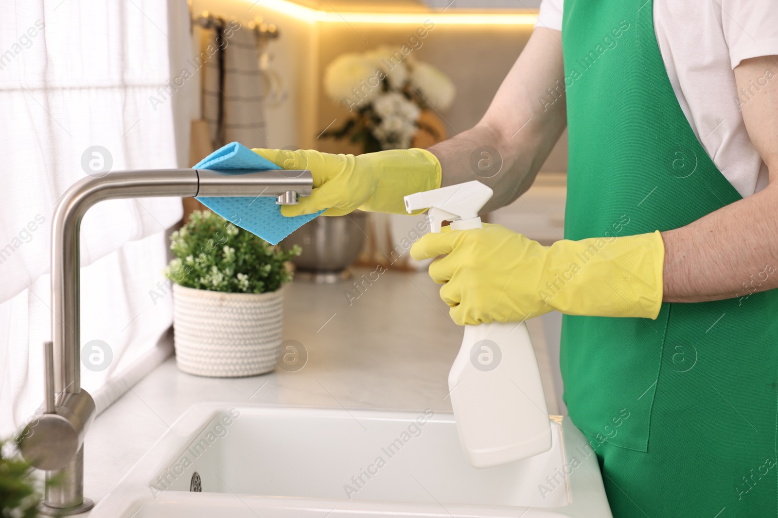 Photo of Professional janitor cleaning tap with rag and detergent in kitchen, closeup