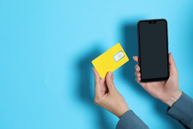 Woman holding SIM card and smartphone on light blue background, closeup. Space for text